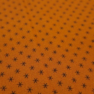 May include: Close-up of orange fabric with a repeating pattern of small, dark blue star-like shapes. The fabric has a slightly textured appearance, with the stars evenly spaced across the surface.