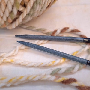 May include: Two blue knitting needles with black tips, lying on a white surface with a ball of white, brown, and yellow yarn.