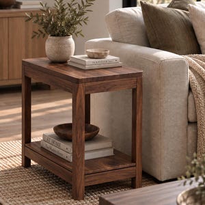 Puede incluir: Una mesa auxiliar rectangular de madera marrón oscuro con cuatro patas rectas y un estante inferior. La superficie superior sostiene un jarrón beige texturizado con vegetación, un cuenco pequeño y dos libros titulados The Kinfolk Home.