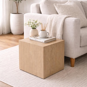 May include: A light oak cube-shaped side table with a natural wood grain finish. The table is in a living room setting, with a sofa, rug, and decorative items on top. The table is 18 inches tall and 18 inches wide.