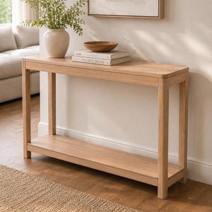 May include: A rectangular light wood console table with a lower shelf, featuring a smooth top surface. On the table sits a beige ceramic vase with green foliage, a small wooden bowl, and two stacked books with the text Live Beautifully.