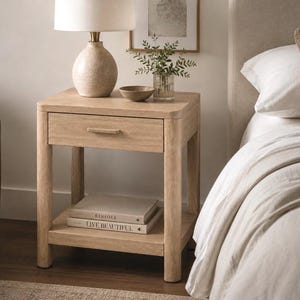 Puede incluir: Una mesita de noche de madera de tono claro con un cajón único con tirador horizontal y un estante inferior abierto. El estante contiene dos libros apilados titulados KINFOLK y LIVE BEAUTIFUL. La mesita tiene esquinas redondeadas y cuatro patas.
