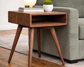 Può includere: Tavolino in stile moderno di metà secolo in legno scuro. Il tavolo ha un piano rettangolare con un ripiano incassato e gambe angolate. Una piccola lampada bianca con base dorata e una pianta in vaso sono sopra. Misura circa 60 cm di lunghezza.