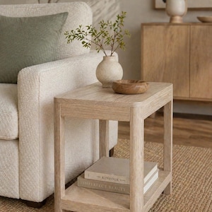 Puede incluir: Una mesa auxiliar de roble claro con un estante inferior que contiene dos libros. Un pequeño jarrón con vegetación y un cuenco de madera están encima. La mesa está junto a un sillón color crema y una alfombra tejida.