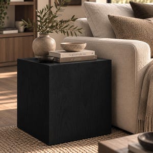 Puede incluir: Una mesa auxiliar cúbica de madera negra con acabado liso y veta visible. Encima hay un jarrón de cerámica beige con ramas verdes, un cuenco pequeño oscuro, un cuenco moteado y dos libros apilados titulados The Kinfolk Home y At Home.