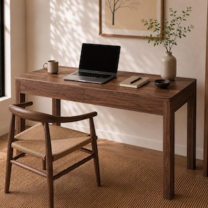 May include: A dark wood desk with two drawers and a matching chair featuring a woven seat. On the desk sits a laptop, a ceramic mug, a notebook with a pen, and a small bowl. A vase with green branches is placed on the desk surface.