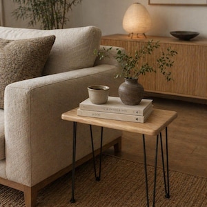 May include: A small wooden side table with black hairpin legs. The table holds two books, a small ceramic cup, and a decorative vase with greenery. A beige sofa and a woven rug are in the background, creating a cozy living room scene.