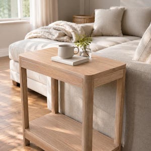 May include: A light-toned wooden side table with a rectangular top and a lower shelf. The table features rounded corners and four straight legs. A white ceramic mug and a small glass vase with green sprigs sit on top of the wooden surface.