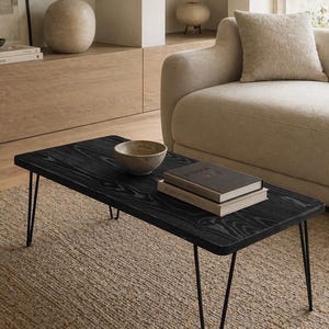 May include: A black rectangular coffee table with metal hairpin legs. The table has a textured wood grain surface. A small bowl and two books are on the table. The table is on a woven rug.