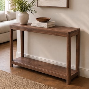 May include: A rectangular dark wood console table with four straight legs and a lower shelf. On top sits a light-colored ceramic vase with green foliage, a small dark wooden bowl, and two stacked books with the text LIVE BEAUTIFULLY on the spine.