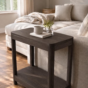 May include: A rectangular dark wood side table with a smooth top surface and a lower shelf. The table features four straight legs and rounded corners on the top panel. A white ceramic mug and a small glass vase with green leaves sit on the table.