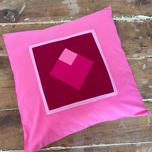 Pu&ograve; includere: Un cuscino rosa con un quadrato rosso al centro. Il quadrato &egrave; composto da quadrati pi&ugrave; piccoli in tonalit&agrave; di rosa.