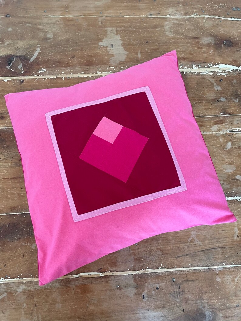 Pu&ograve; includere: Un cuscino rosa con un quadrato rosso al centro. Il quadrato &egrave; composto da quadrati pi&ugrave; piccoli in tonalit&agrave; di rosa.