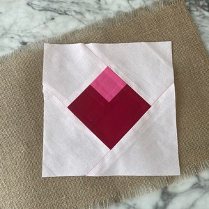 Pu&ograve; includere: Un blocco di trapunta rosa e rosso con una forma di diamante al centro. Il blocco &egrave; composto da quadrati di tessuto cuciti insieme.