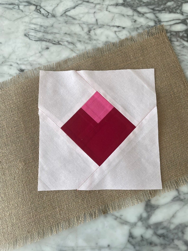 Pu&ograve; includere: Un blocco di trapunta rosa e rosso con una forma di diamante al centro. Il blocco &egrave; composto da quadrati di tessuto cuciti insieme.