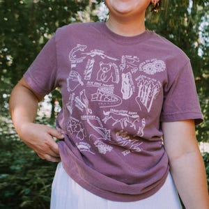 May include: A mauve short-sleeved t-shirt with a white graphic design featuring various illustrations. The person is wearing a white skirt and glasses. The background is a lush green forest.