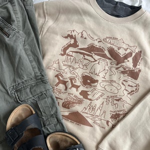 May include: A tan sweatshirt with a brown graphic print of a map of Idaho. The map includes the names of various landmarks and natural features, such as the Snake River, Shoshone Falls, and the Tetons. The sweatshirt is paired with a pair of green cargo pants and black sandals.