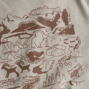 May include: A beige t-shirt with a brown illustration of a map of Idaho. The map features various landmarks and natural features, including the Snake River, Tetons, Sawtooth Mountains, Shoshone Falls, and Coeur d'Alene Lake. The text "Idaho" is printed in the center of the map.