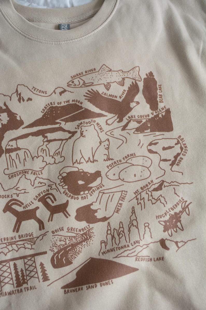 May include: A beige t-shirt with a brown illustration of a map of Idaho. The map features various landmarks and natural features, including the Snake River, Tetons, Sawtooth Mountains, Shoshone Falls, and Coeur d'Alene Lake. The text "Idaho" is printed in the center of the map.