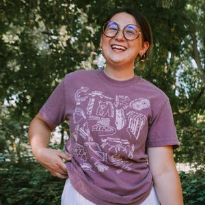 May include: A woman wearing a purple t-shirt with a white graphic design of various animals and plants. The graphic includes the text "Blue Ridge Parkway" and "Mountain to Sea Trail". She is wearing a white skirt and smiling.