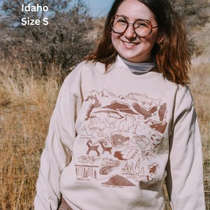 May include: A beige sweatshirt with a brown graphic print of an Idaho state map featuring various landmarks and animals. The text "Idaho" and "Size S" is printed on the top left corner of the sweatshirt.