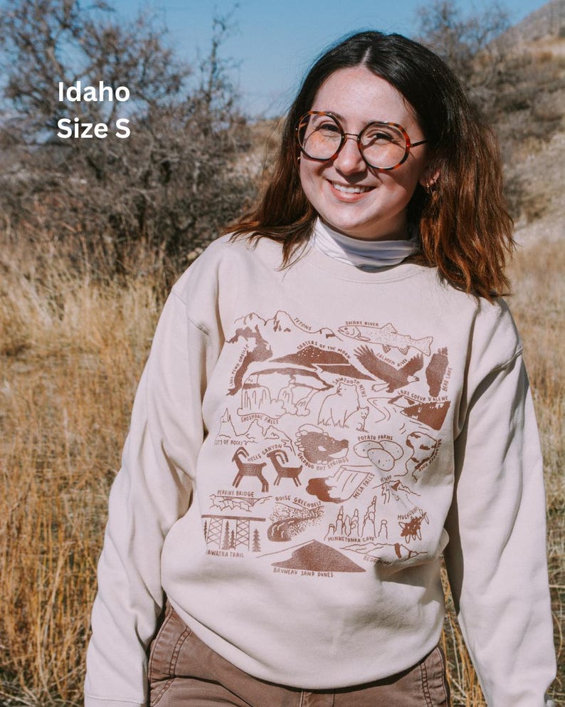 May include: A beige sweatshirt with a brown graphic print of an Idaho state map featuring various landmarks and animals. The text "Idaho" and "Size S" is printed on the top left corner of the sweatshirt.