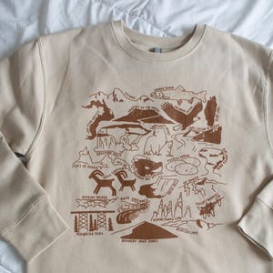 May include: A tan sweatshirt with a brown graphic print of a map of Idaho. The map features various landmarks and natural features, including the Tetons, the Snake River, the Sawtooth Mountains, and the Bruneau Sand Dunes.