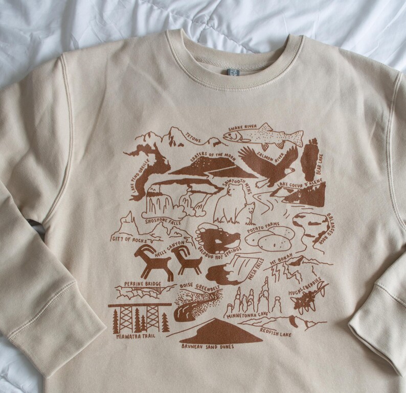 May include: A tan sweatshirt with a brown graphic print of a map of Idaho. The map features various landmarks and natural features, including the Tetons, the Snake River, the Sawtooth Mountains, and the Bruneau Sand Dunes.