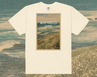 May include: Cream-colored t-shirt featuring a rectangular graphic with a coastal scene. The graphic displays the text "SAN DIEGO CALIFORNIA" above a depiction of the ocean and a grassy hillside. The shirt has short sleeves and a crew neck.