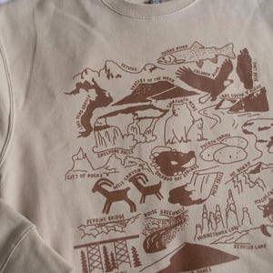 May include: A beige sweatshirt with a brown graphic print of a map of Idaho. The map features various landmarks and natural features, including the Tetons, Crater of the Moon, Shoshone Falls, and the Boise Greenbelt.