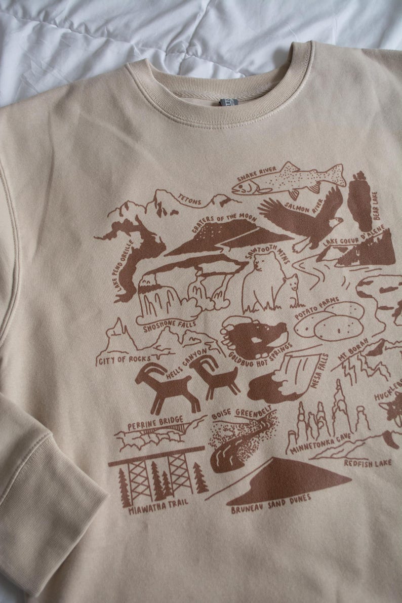 May include: A beige sweatshirt with a brown graphic print of a map of Idaho. The map features various landmarks and natural features, including the Tetons, Crater of the Moon, Shoshone Falls, and the Boise Greenbelt.