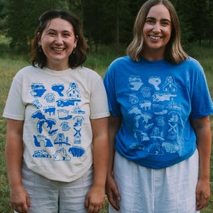May include: Two people wearing white t-shirts with blue illustrations of landmarks and places. The person on the left is wearing a cream colored t-shirt and the person on the right is wearing a royal blue t-shirt. Both shirts feature a variety of illustrations, including a lighthouse, a bridge, a windmill, and a lake.