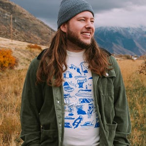 May include: A man wearing a green corduroy jacket over a white t-shirt with a blue graphic print of various landmarks. He is wearing a gray beanie and has long brown hair. He is standing in a field with mountains in the background.