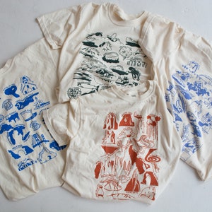 May include: Four cream-colored t-shirts with different blue, green, and orange graphic prints of various landmarks. The prints are stylized and resemble illustrations.
