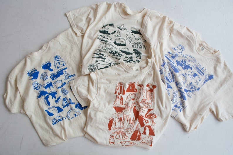 May include: Four cream-colored t-shirts with different blue, green, and orange graphic prints of various landmarks. The prints are stylized and resemble illustrations.