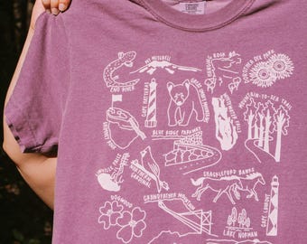 May include: Purple t-shirt with a white graphic design featuring various landmarks and symbols from North Carolina. The design includes the words "MT Mitchell", "ENO River", "Blue Ridge Parkway", "Grandfather Mountain", "Shackleford Banks", "Pilot Mountain", "Bald Head Island", "Black Balsam Knob", "Cape Hatteras", "Hanging Rock", "Great Smoky Mountains", "Whitewater Falls", "Mountain-to-Sea Trail", "Dogwood", "Northern Cardinal", "Lake Norman", "Cape Lookout", "Dorothea Dix Park".