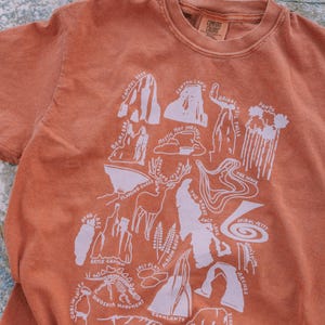 May include: A rust-colored t-shirt with a white graphic design of a map of national parks in Utah. The map includes locations like Zion National Park, Arches National Park, Canyonlands National Park, Bryce Canyon National Park, Capitol Reef National Park, and Goblin Valley State Park.