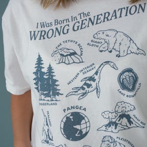 May include: White t-shirt with a blue graphic print that says "I Was Born In The Wrong Generation". The graphic includes illustrations of extinct animals and geological formations, including a giant sloth, a plesiosaur, a trilobite, and a volcano.