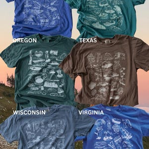 May include: Five t-shirts with different state names and illustrations of landmarks from each state. The states are Minnesota, Colorado, Oregon, Texas, and Virginia. The shirts are all different colors, including blue, green, brown, and gray.