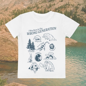 May include: White t-shirt with the text "I Was Born In The Wrong Generation" and illustrations of prehistoric animals and geological features in navy blue. The shirt has a crew neck and short sleeves.