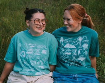 May include: Two people wearing teal blue t-shirts with white outlines of various landmarks and natural features. The t-shirts have the text "SEQUOIA AND KINGS CANYON NATIONAL PARKS" on the front. The people are sitting in a grassy field.