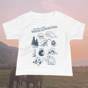 May include: White t-shirt with a blue graphic print that says "I Was Born In The WRONG GENERATION". The graphic features a circle with illustrations of different prehistoric animals and geological formations, including a giant sloth, a mammoth, a trilobite, and a volcano.