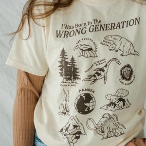 May include: A cream-colored t-shirt with a brown graphic print that says "I Was Born In The Wrong Generation". The graphic includes illustrations of extinct animals and geological formations, including a giant sloth, a sea serpent, a trilobite, a volcano, a glacier, and a mammoth.