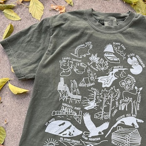 May include: A green t-shirt with a white graphic design featuring a map of Pennsylvania with various landmarks and locations. The text on the shirt includes "Susquehanna River", "Laurel Caverns", "Mt. Minsi", "The Pogonip", "Ohiopyle", "Black Moshannon", "Mountain Laurel", "Lehigh Gorge", "Presque Isle Bay", "Allegheny Nat'l Forest", "Ricketts Glen", "Cherry Springs", "Bushkill Falls", "Schuylkill River", "PA Grand Canyon", "Hawk Mountain", "Valley Forge", "Delaware Water Gap", and "Longwood Gardens".