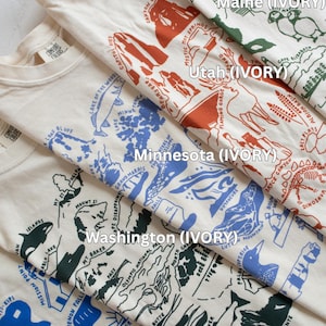 May include: Five different t-shirts with state-themed illustrations and the text "(IVORY)" printed on each. The states featured are Maine, Utah, Minnesota, Washington, and Michigan.