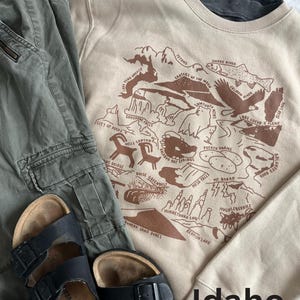 Puede incluir: Una sudadera beige con un estampado gr&aacute;fico marr&oacute;n de un mapa de Idaho. El mapa presenta varios puntos de referencia e inter&eacute;s, incluyendo el r&iacute;o Snake, los Tetons y el Monumento Nacional del Cr&aacute;ter de la Luna. El texto "Idaho" est&aacute; impreso en marr&oacute;n en la parte inferior de la sudadera.