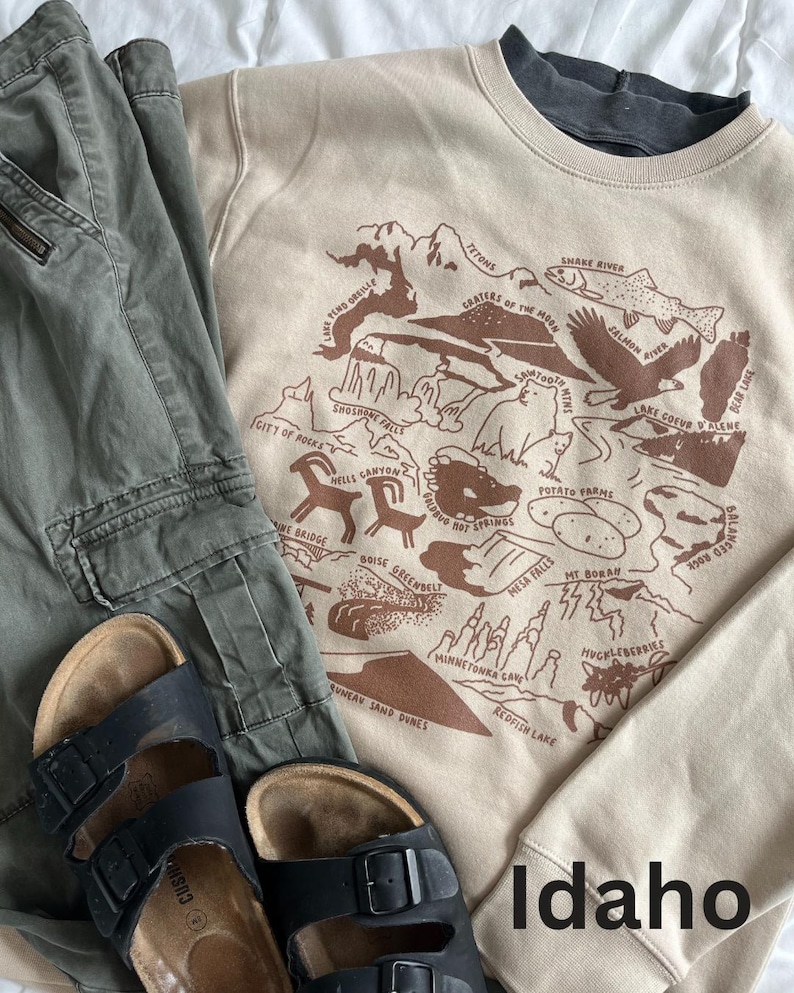 Puede incluir: Una sudadera beige con un estampado gr&aacute;fico marr&oacute;n de un mapa de Idaho. El mapa presenta varios puntos de referencia e inter&eacute;s, incluyendo el r&iacute;o Snake, los Tetons y el Monumento Nacional del Cr&aacute;ter de la Luna. El texto "Idaho" est&aacute; impreso en marr&oacute;n en la parte inferior de la sudadera.
