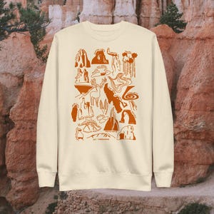 May include: A cream-colored sweatshirt with an orange graphic print of a map of Arches National Park in Utah. The map includes landmarks like Delicate Arch, Balanced Rock, and Double Arch. The text "MT. TIMPANOGOS" is printed at the bottom of the map.