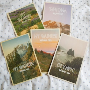 May include: A collection of five vintage-style postcards featuring scenic views of national parks. The postcards display images of mountains, forests, and coastlines, with text that reads "MT RAINIER NATIONAL PARK", "TAHOMA WASHINGTON", and "OLYMPIC NATIONAL PARK".