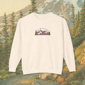 以下が含まれることがあります： クリーム色のスウェットシャツ。紫色のアーチの中に、Mt. Rainier、ヒマワリ、そして「MT. RAINIER」の文字が描かれたグラフィックが特徴です。デザインは胸の中央に配置されています。背景には木々のある山の風景が描かれています。